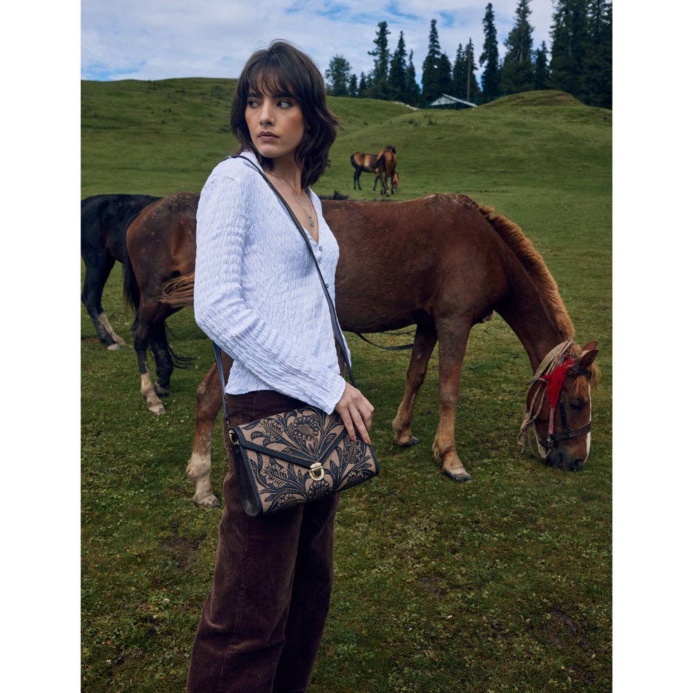 Woman standing in a grassy field with horses, holding a patterned bag.