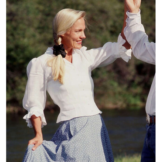 Lady wearing a white Ladies Angel Blouse with ruffled sleeves, smiling outdoors, paired with a blue skirt.
