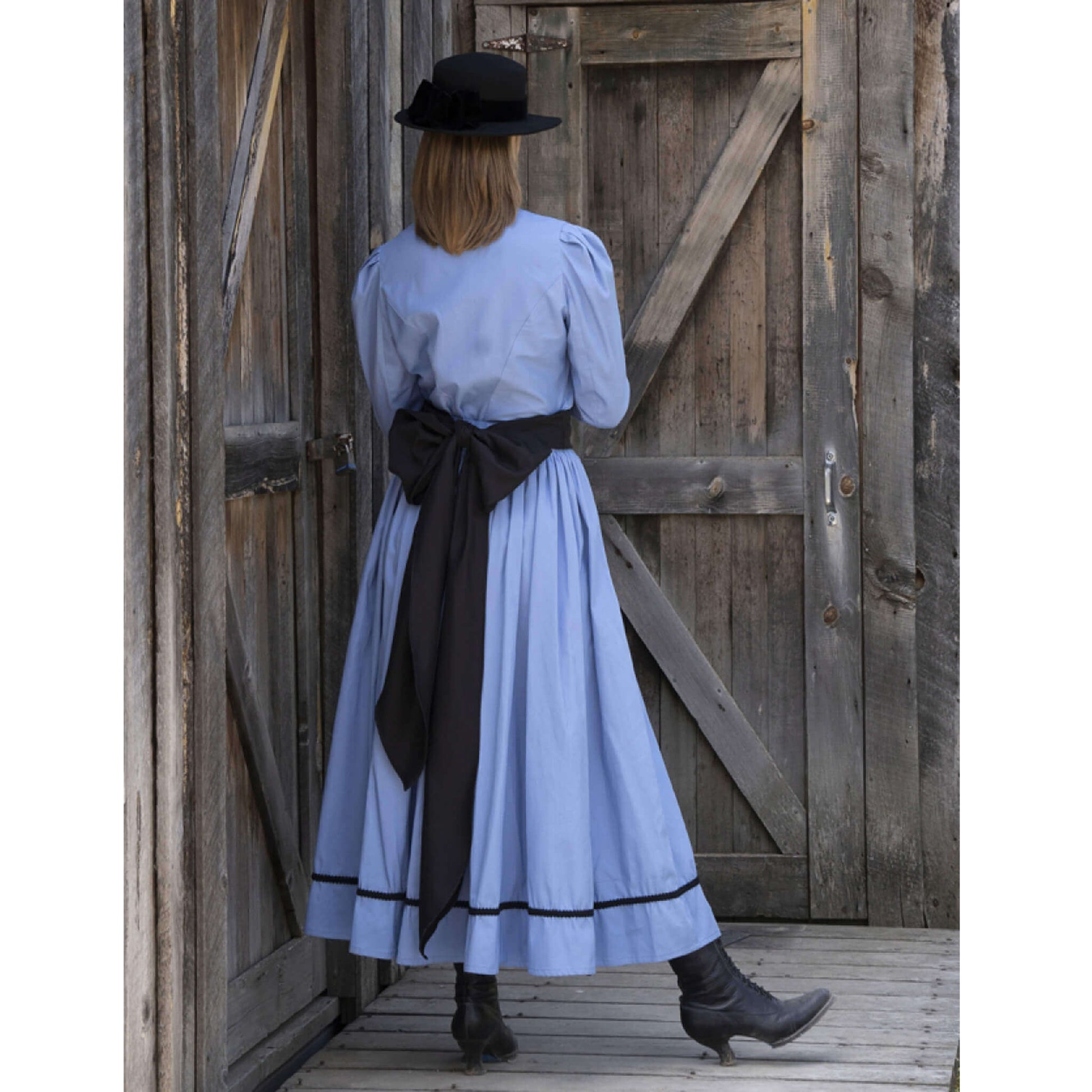 Back view of a woman in a Ladies Old West Bowman Outfit with a blue dress and black accents, standing near wooden doors.