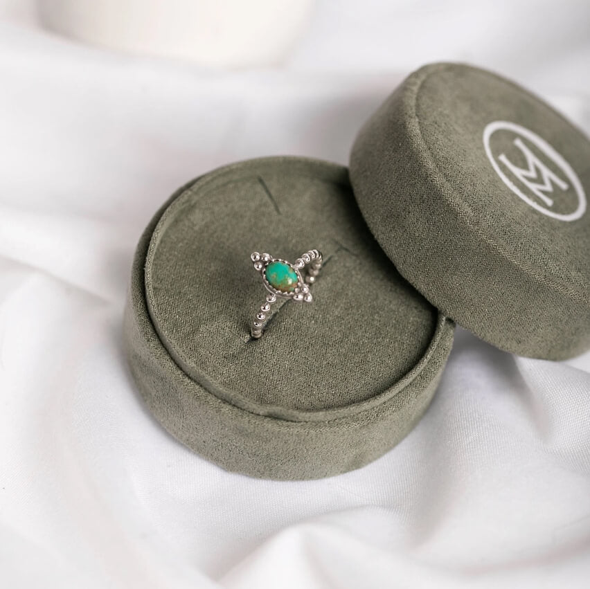 A silver ring with a green oval center stone and small clear stones in a star-like pattern, displayed on a round olive green velvet ring box with its lid beside it on a white fabric background.