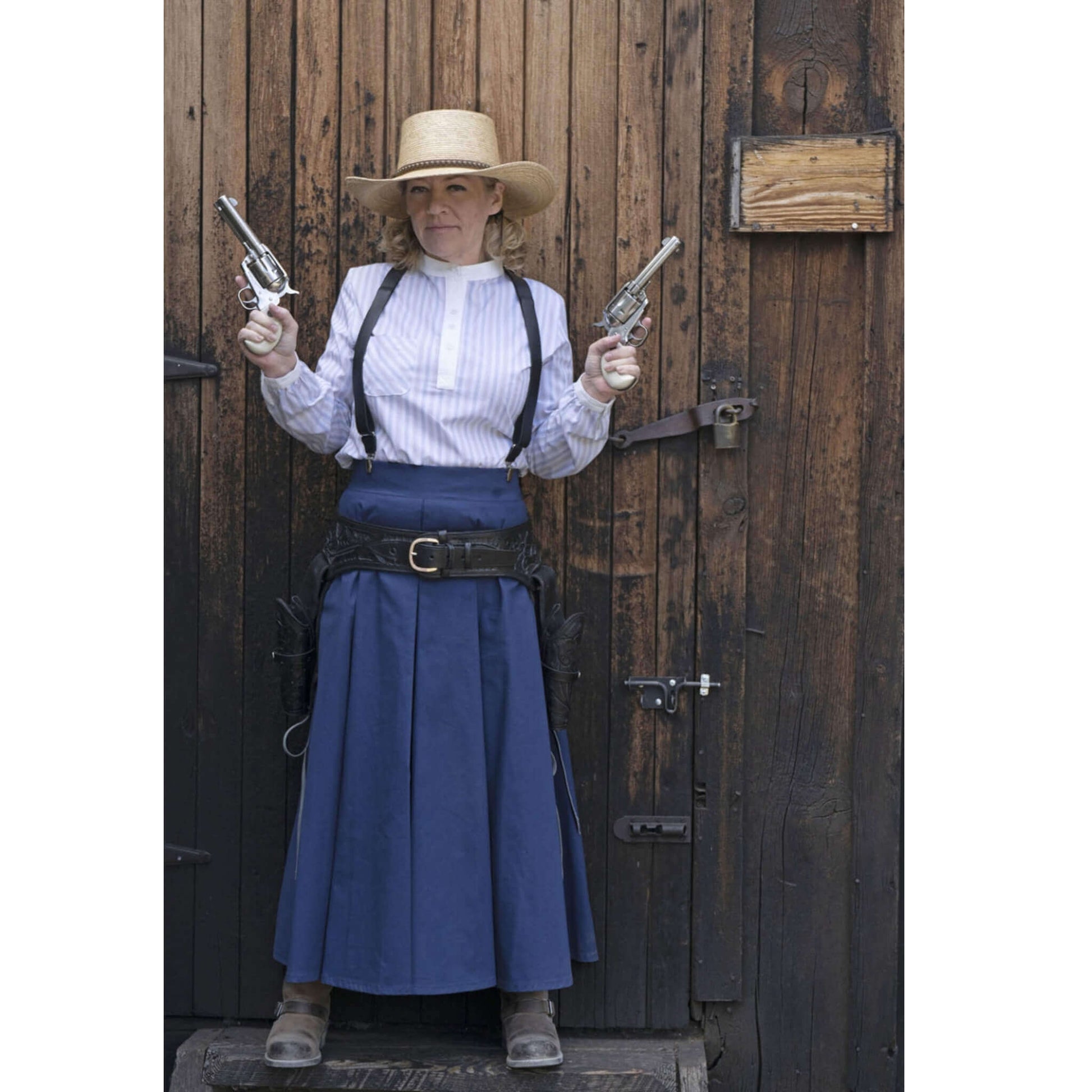 Woman in Old West attire holding revolvers, wearing a traditional blue skirt and shirt, posed against a wooden backdrop.