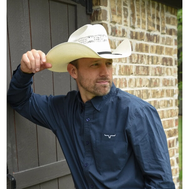 Man wearing a navy blue shirt with a logo, holding a beige cowboy hat against a brick wall.