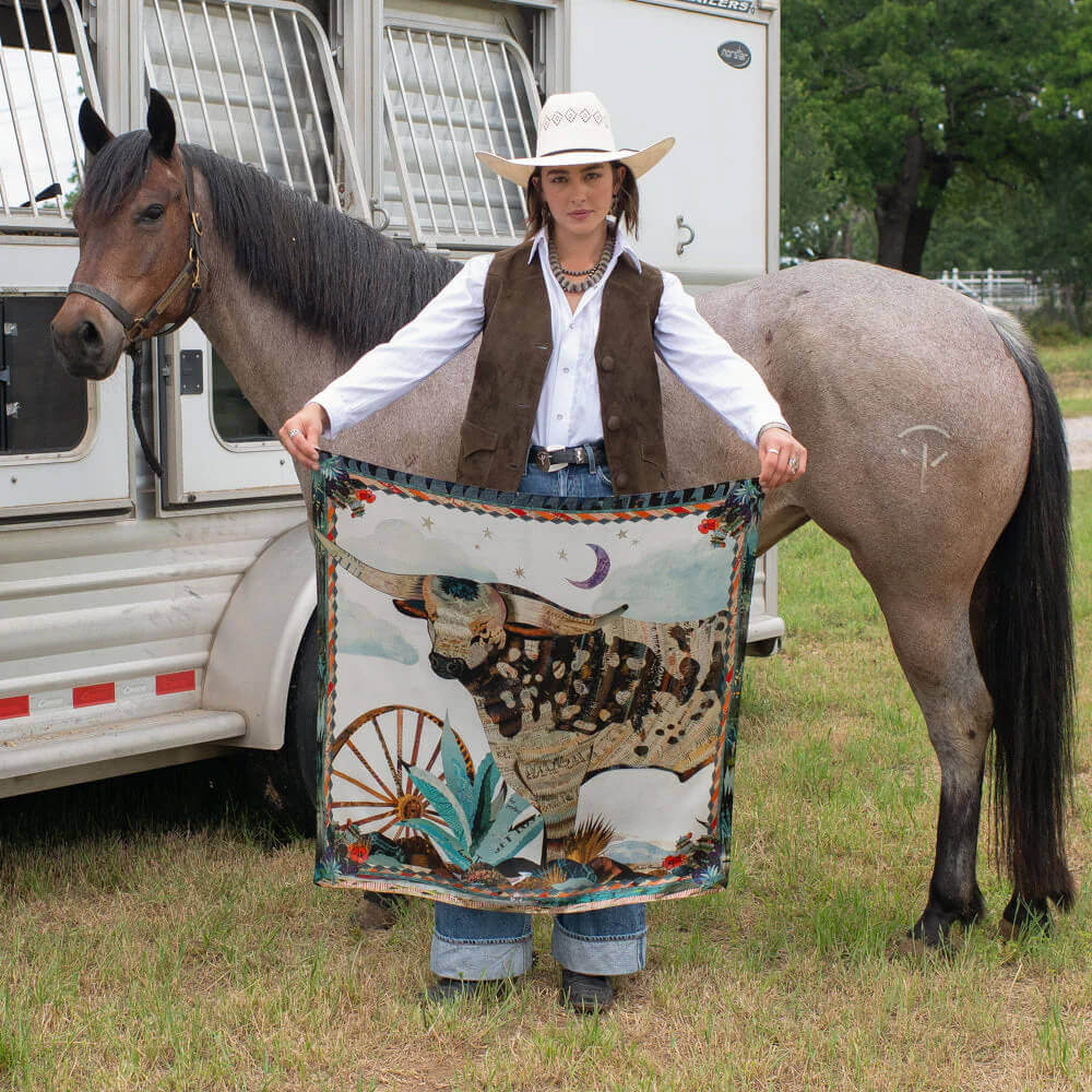 Woman holding the Broken Spoke Western Silk Wild Rag with longhorn steer design next to a horse in a western setting.