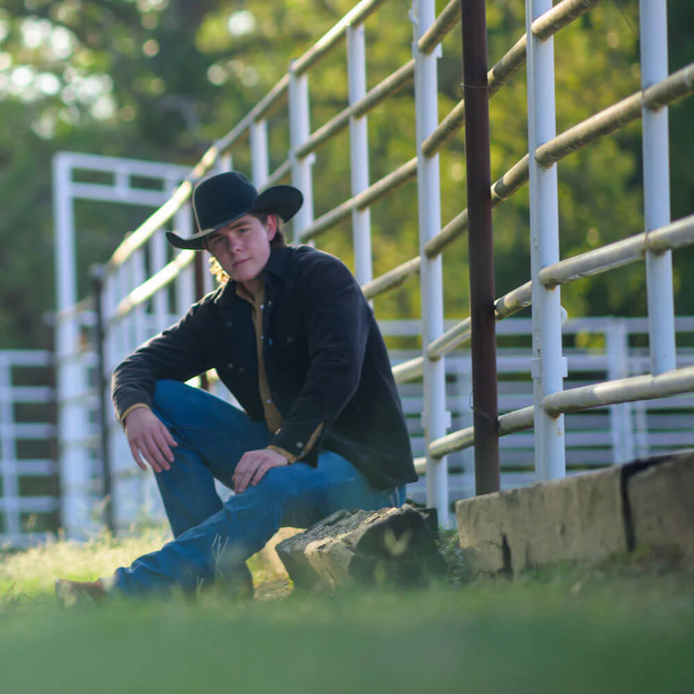 Man in cowboy hat and jacket sitting on a rock in front of a white fence with greenery.