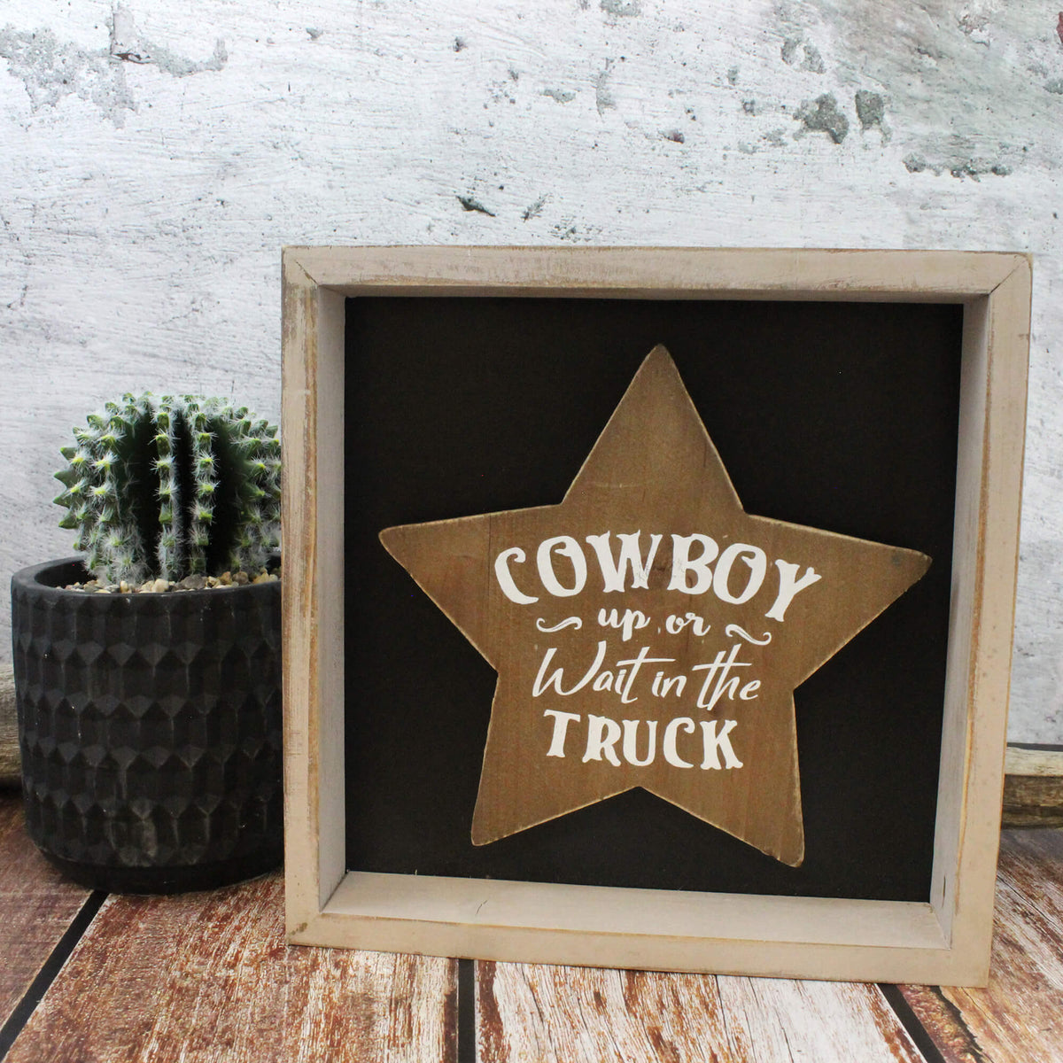 A rustic framed sign featuring a wooden star with the phrase 'COWBOY up or Wait in the TRUCK' in white lettering on a black background, styled on a wooden surface next to a small potted cactus against a distressed white wall.