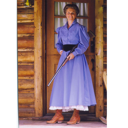 Woman in a blue Old West Farmstead Dress with black cummerbund, standing by a log cabin door.