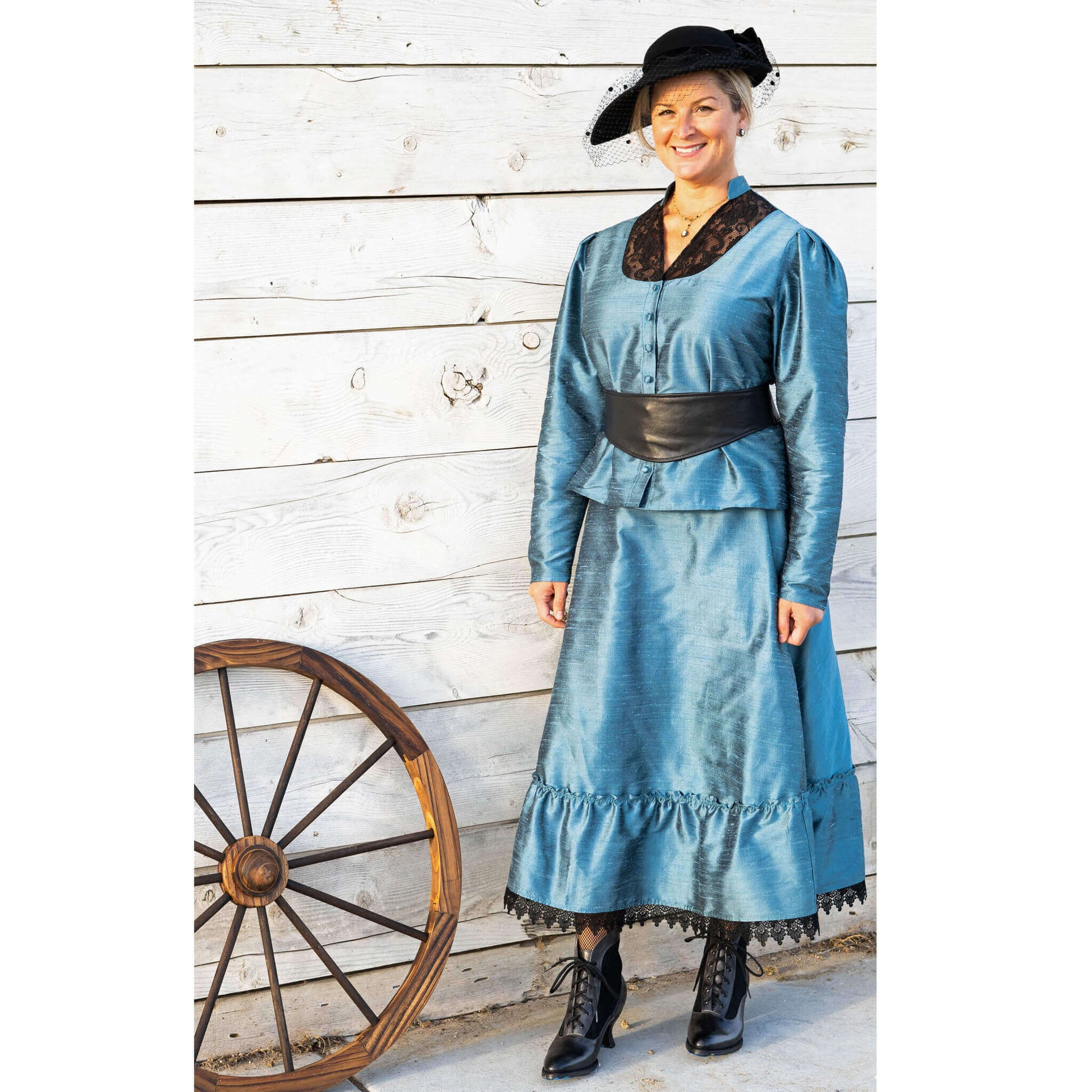 Woman in teal 1800's Old West outfit with black lace accents and hat, next to a wooden wagon wheel.