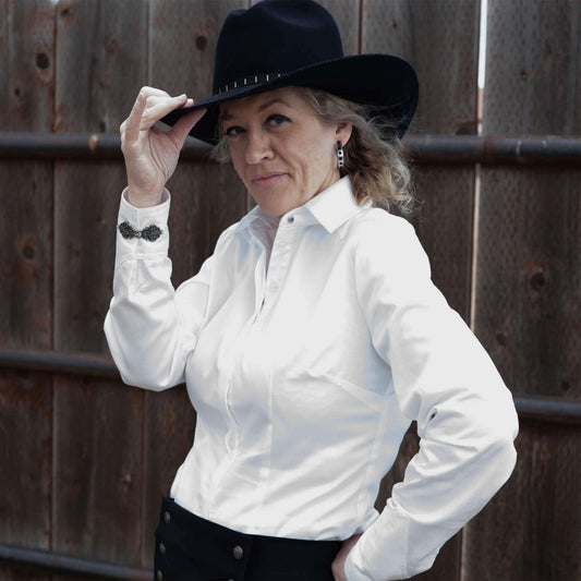 Model wearing a white Western Riding Blouse with unique cuff details and a black cowboy hat, against a rustic wooden background.
