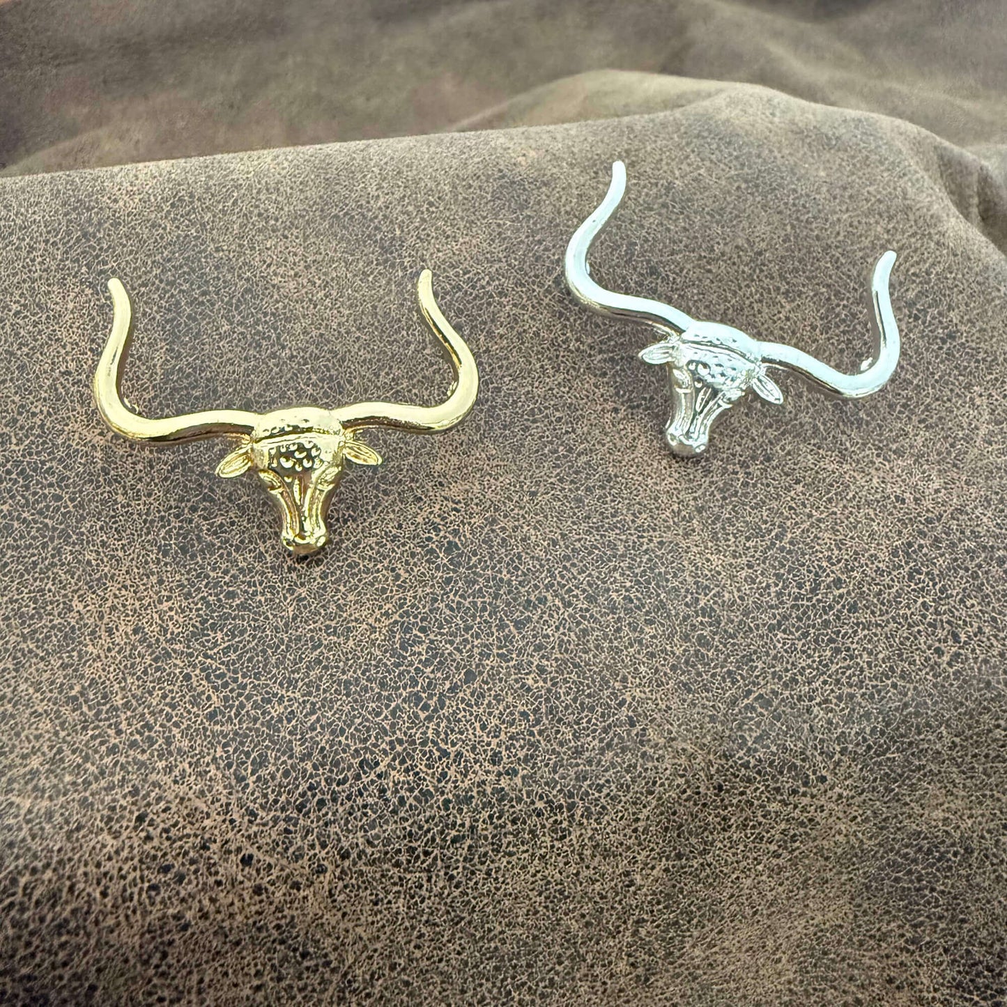 Gold and silver bull skull earrings on a textured brown surface