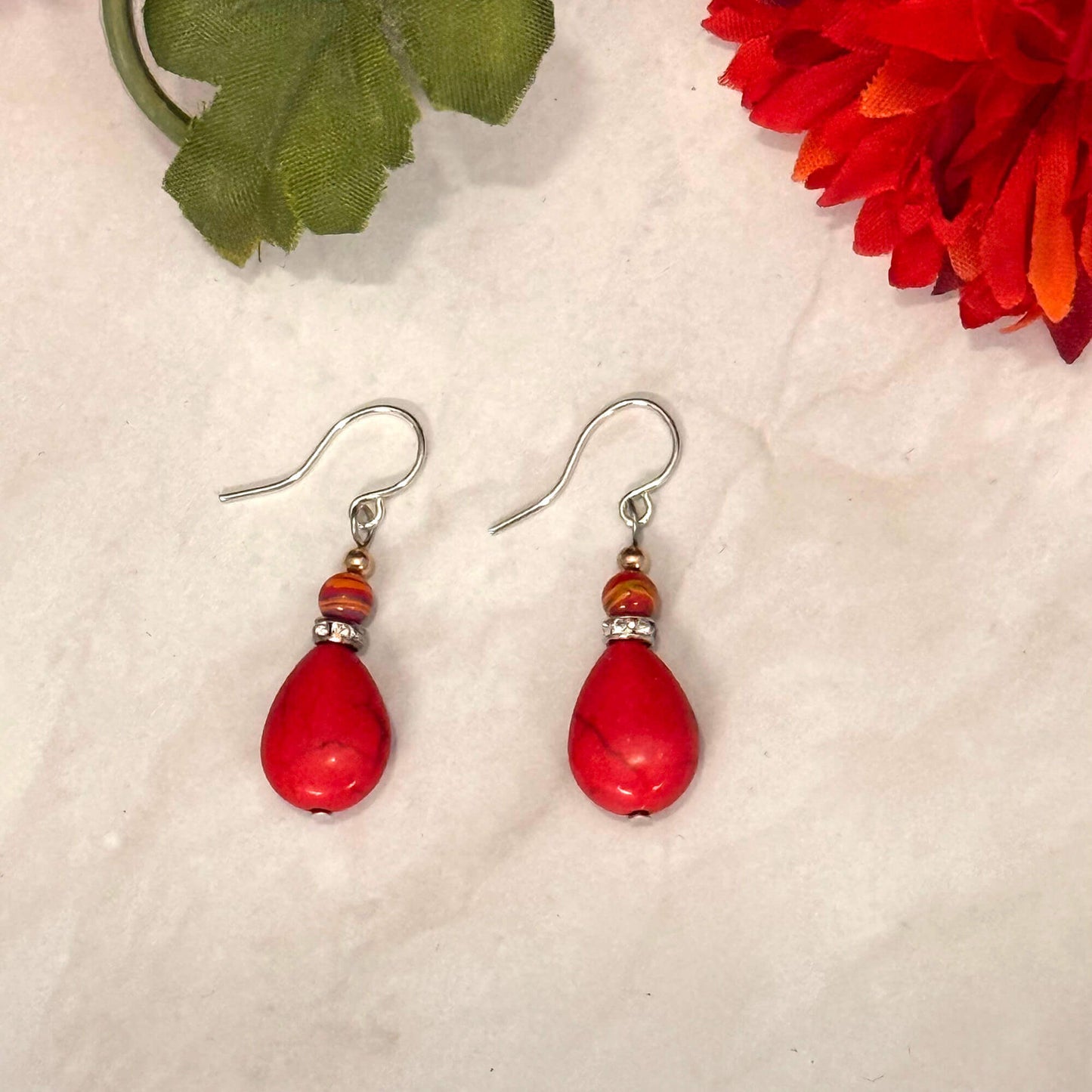 Red teardrop earrings with silver hooks on a textured surface with a red flower and green leaf in the background.