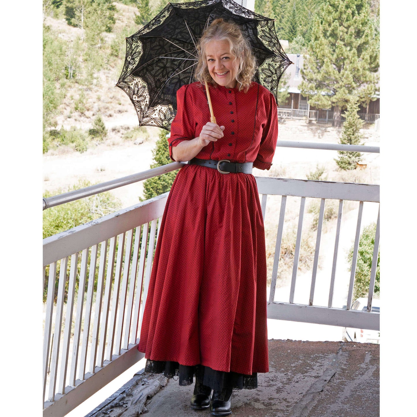 Woman in a red Old West saloon dress holding a black parasol, standing on a balcony with scenic background.