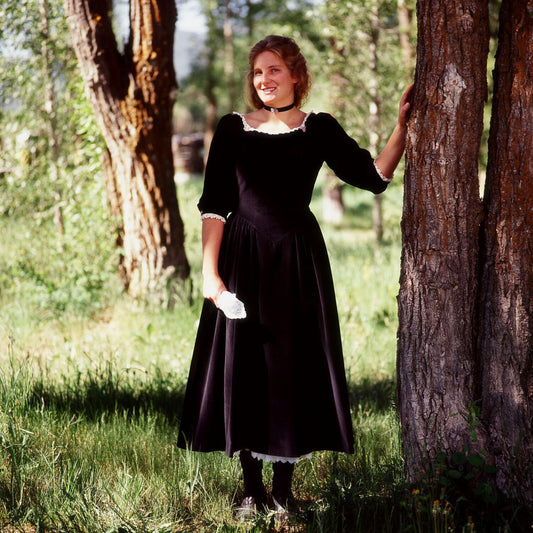 Woman in black velvet Maverick Queen vintage dress standing by a tree in a green forest setting.