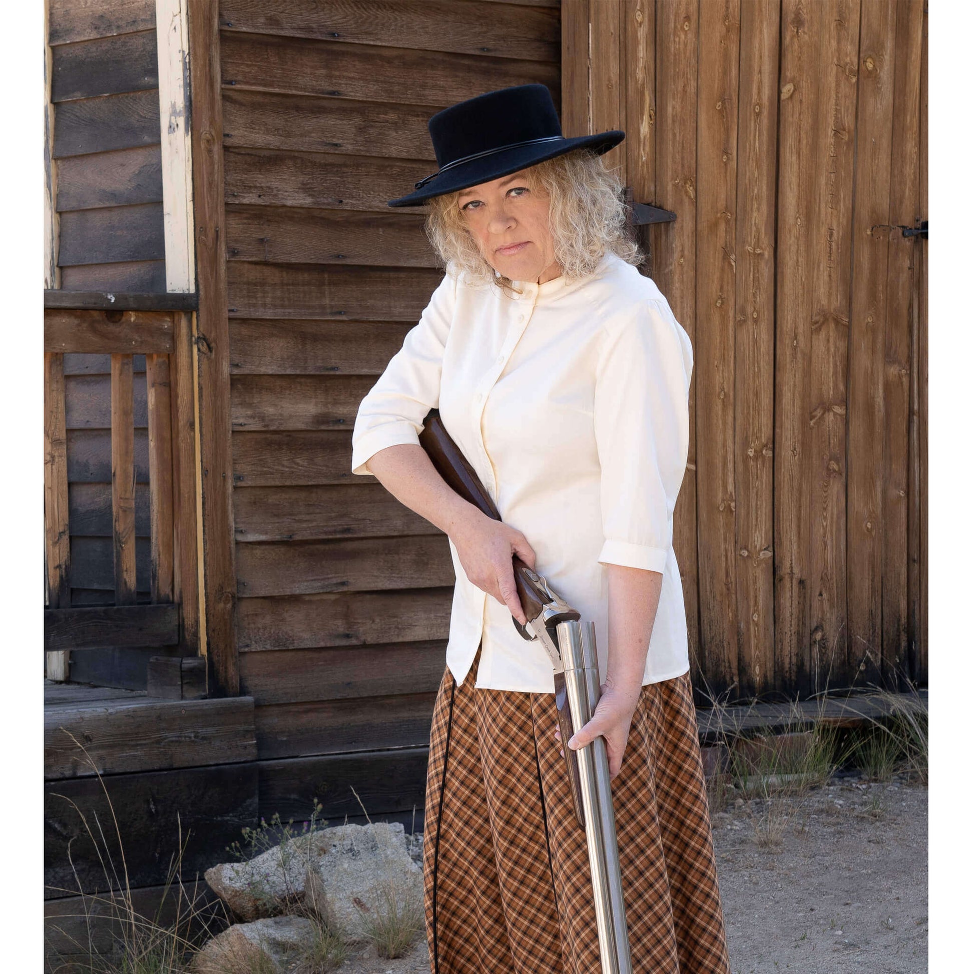 Woman wearing a white western shirt and black hat, posing with a gun in front of a rustic wooden building.