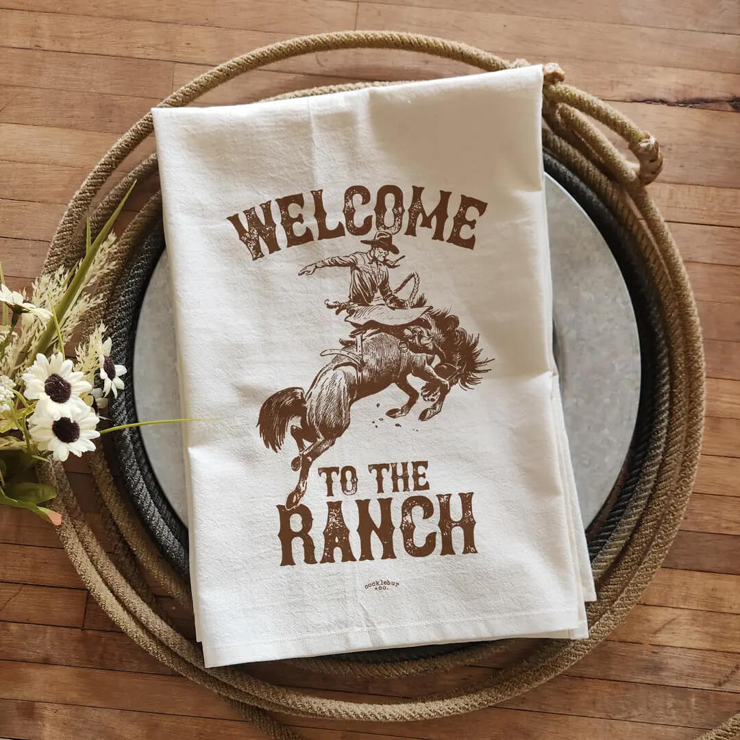 A white fabric tea towel with a brown western-style graphic reading 'WELCOME TO THE RANCH' and showing a cowboy riding a bucking horse, styled on a round tray over a wooden surface with rope and flowers.