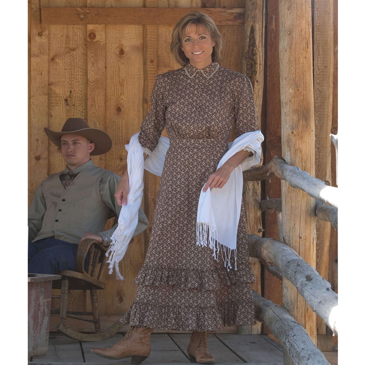Woman wearing Prairie Schooner Authentic Ladies Dress with lace trim, standing in rustic setting next to a man in cowboy attire.