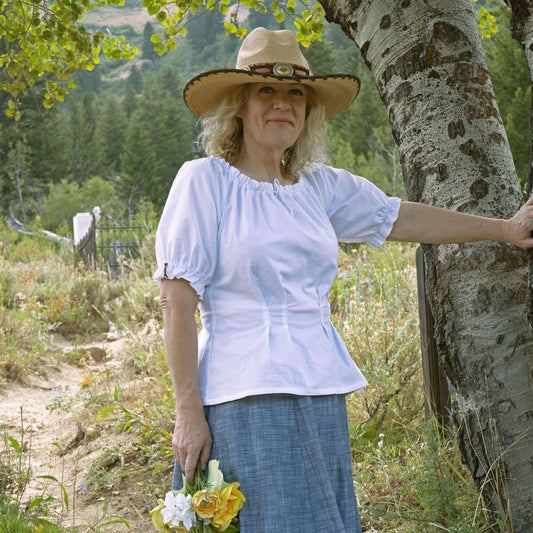 Woman wearing Olmstead Women's Western Peasant Blouse with a hat, holding flowers, in a scenic outdoor setting.