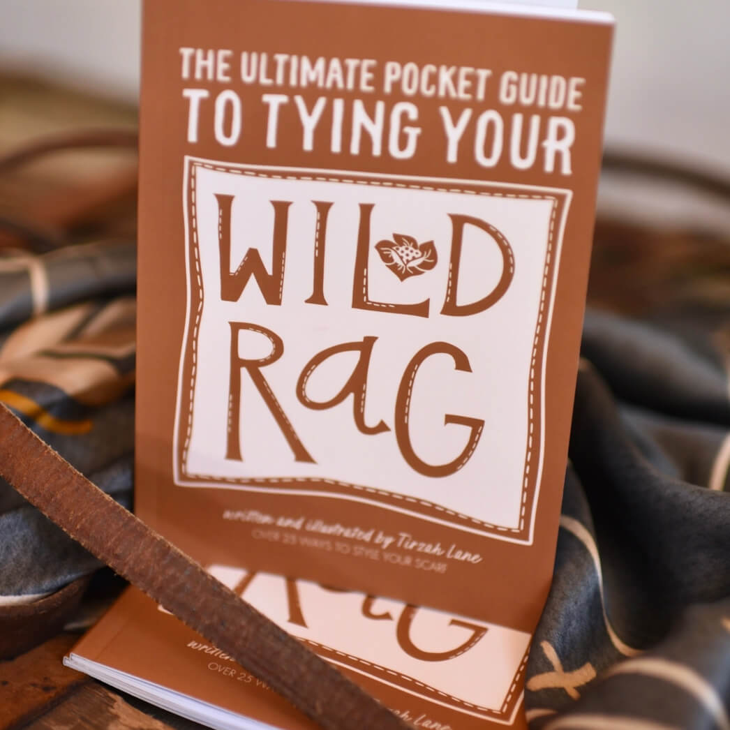 A close-up of a brown pocket guide book titled 'The Ultimate Pocket Guide to Tying Your Wild Rag' resting on a patterned textile with a leather strap in the foreground.