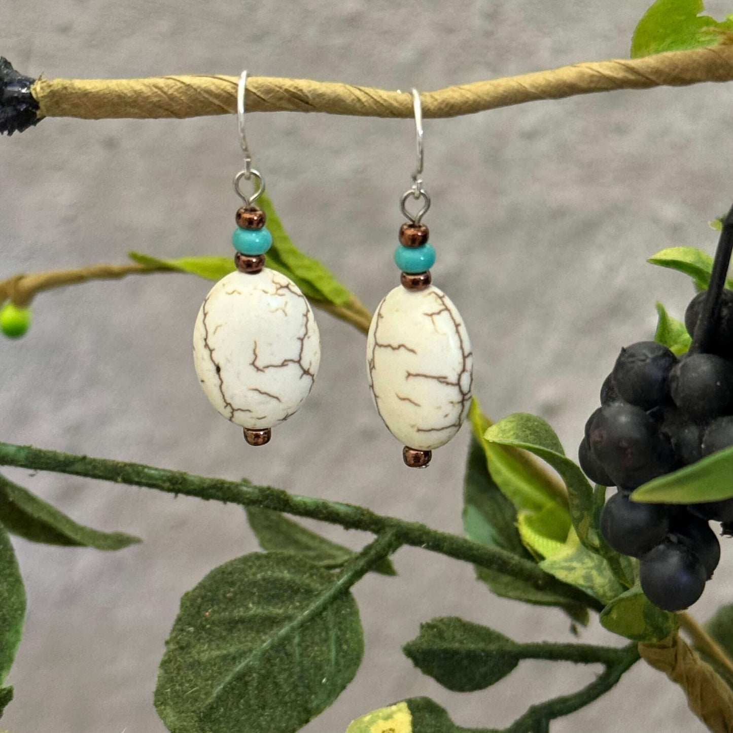 Pair of earrings with white stones and turquoise beads on a branch with leaves and berries.