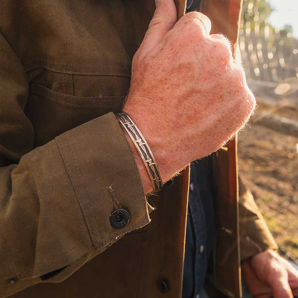 Close-up of a person wearing a brown jacket and bracelet, with a blurred natural background