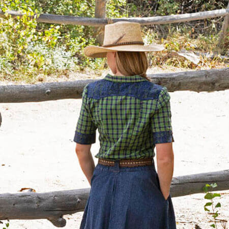 Back view of a woman wearing a Rodeo Round-Up Ladies Shirt with plaid pattern and lace detail, styled with a straw hat.