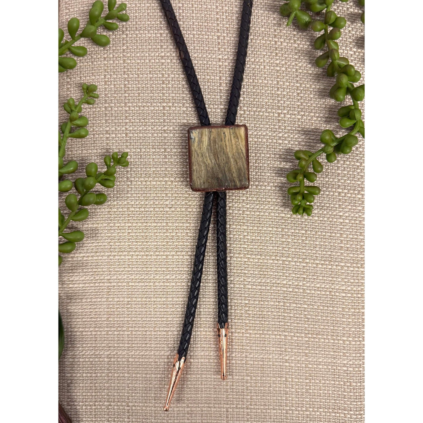 Bolo tie with square wooden pendant on a beige fabric background with green leaves