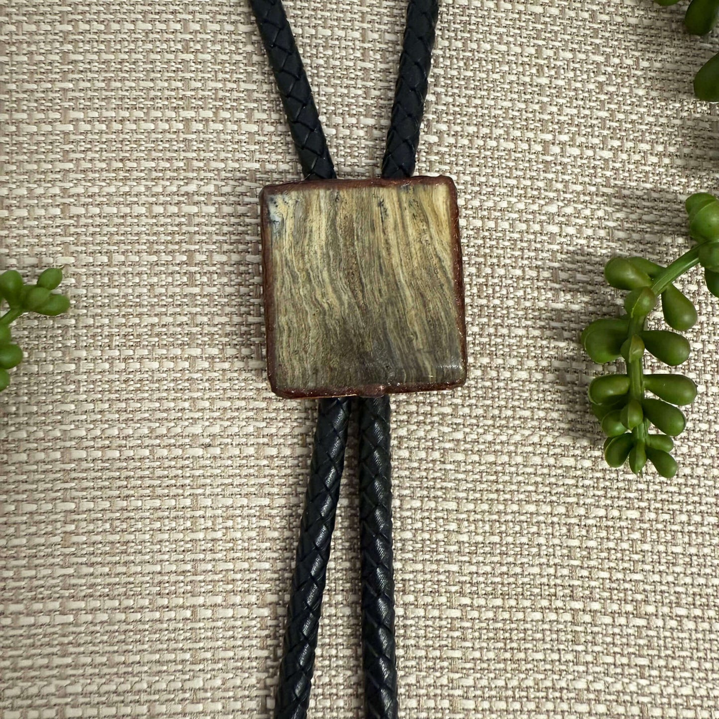 Bolo tie with a square, textured pendant on a textured fabric background with green leaves.