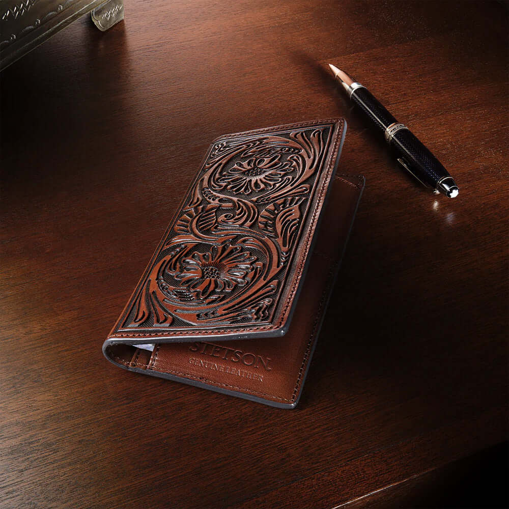 Brown leather wallet with intricate designs on a wooden surface, accompanied by a pen.