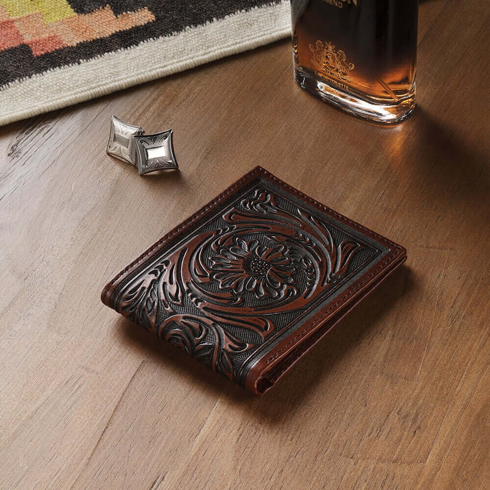 Decorative leather wallet with intricate designs on a wooden surface, next to a glass of whiskey and cufflinks.