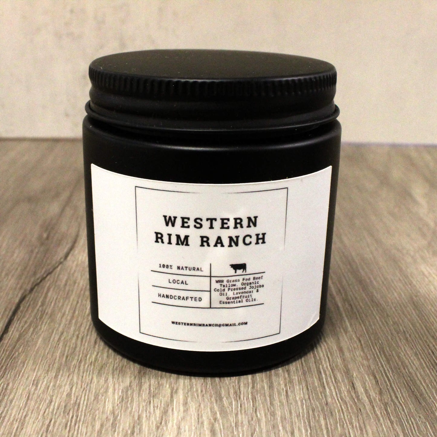 A black glass jar candle with a black screw-on lid and a white paper label reading 'WESTERN RIM RANCH' on a wood surface against a light wall.