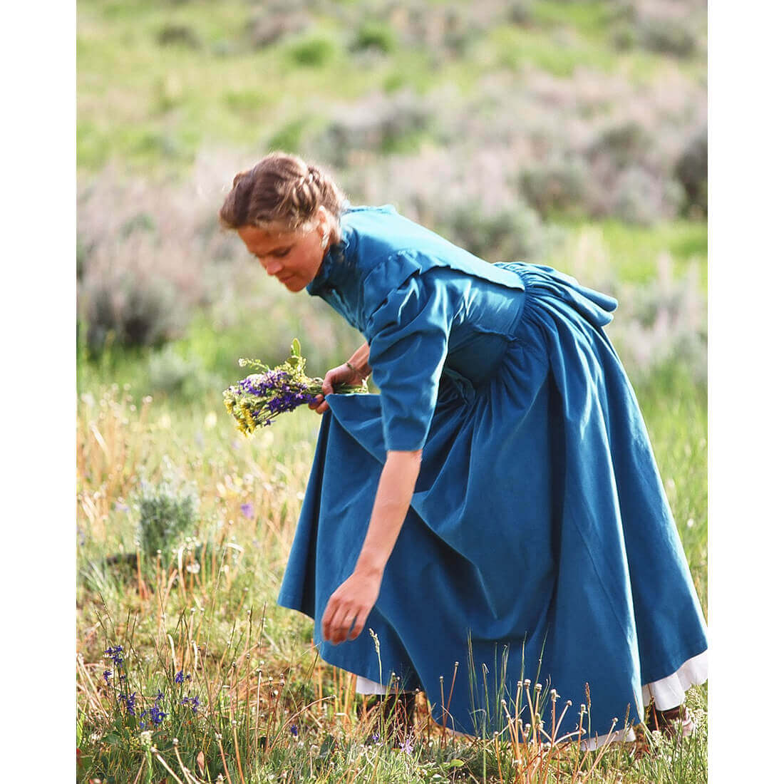 Woman in Prairie Bustle Old West Dress picking wildflowers in a grassy field, showcasing the dress's vintage style.