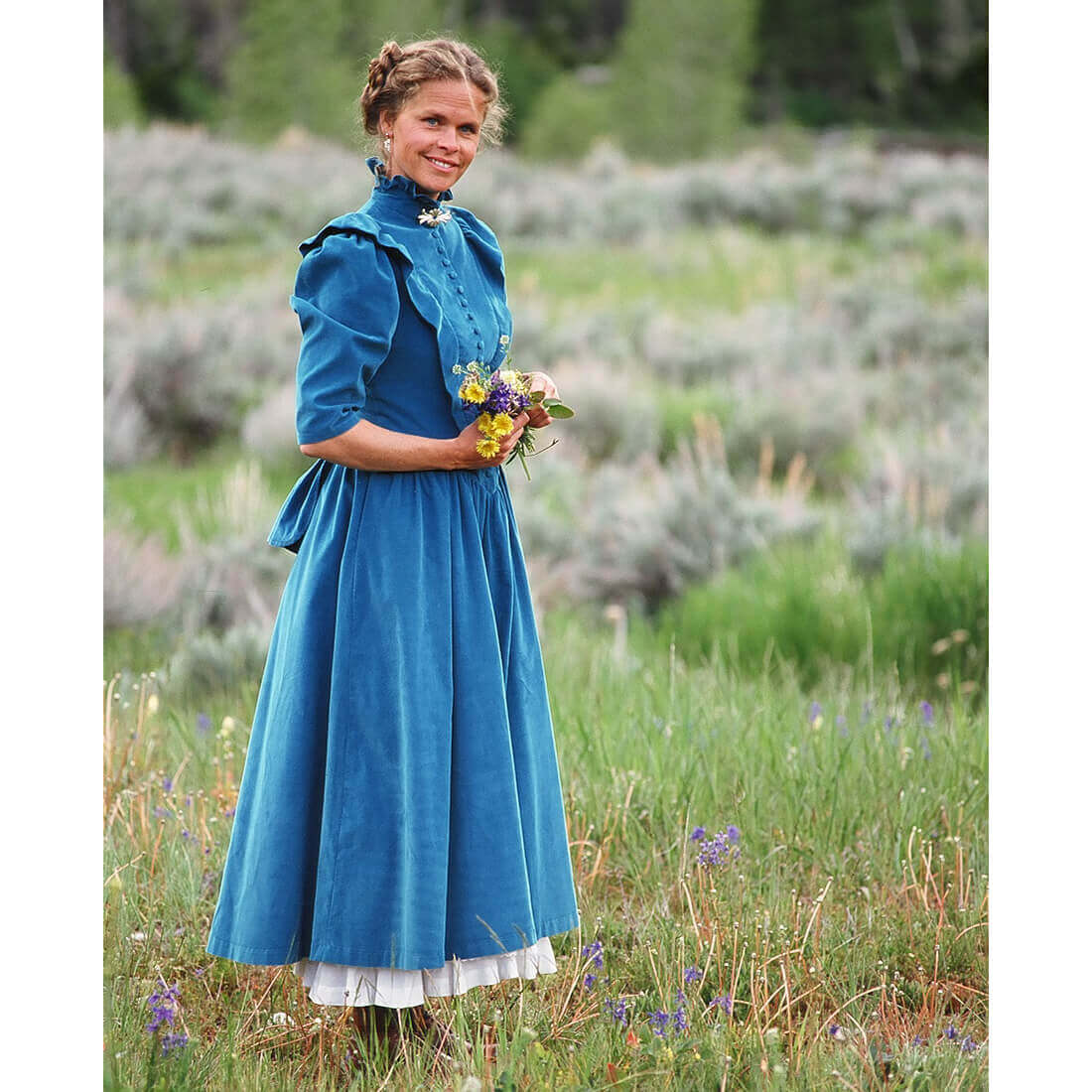 Woman in a Prairie Bustle Old West Dress holding flowers in a scenic field with greenery and wildflowers.