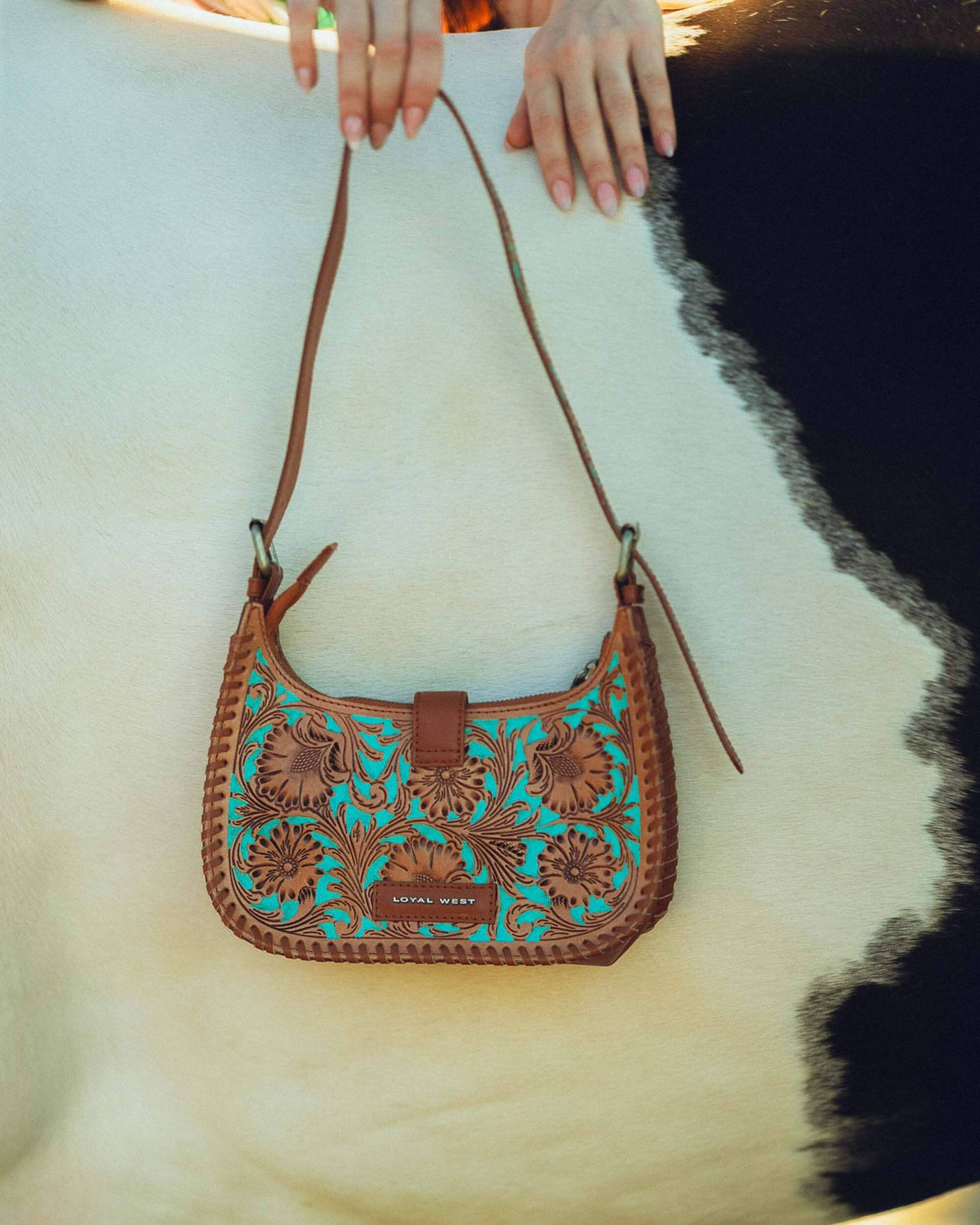 Brown leather handbag with teal embroidery held by a person on a white background