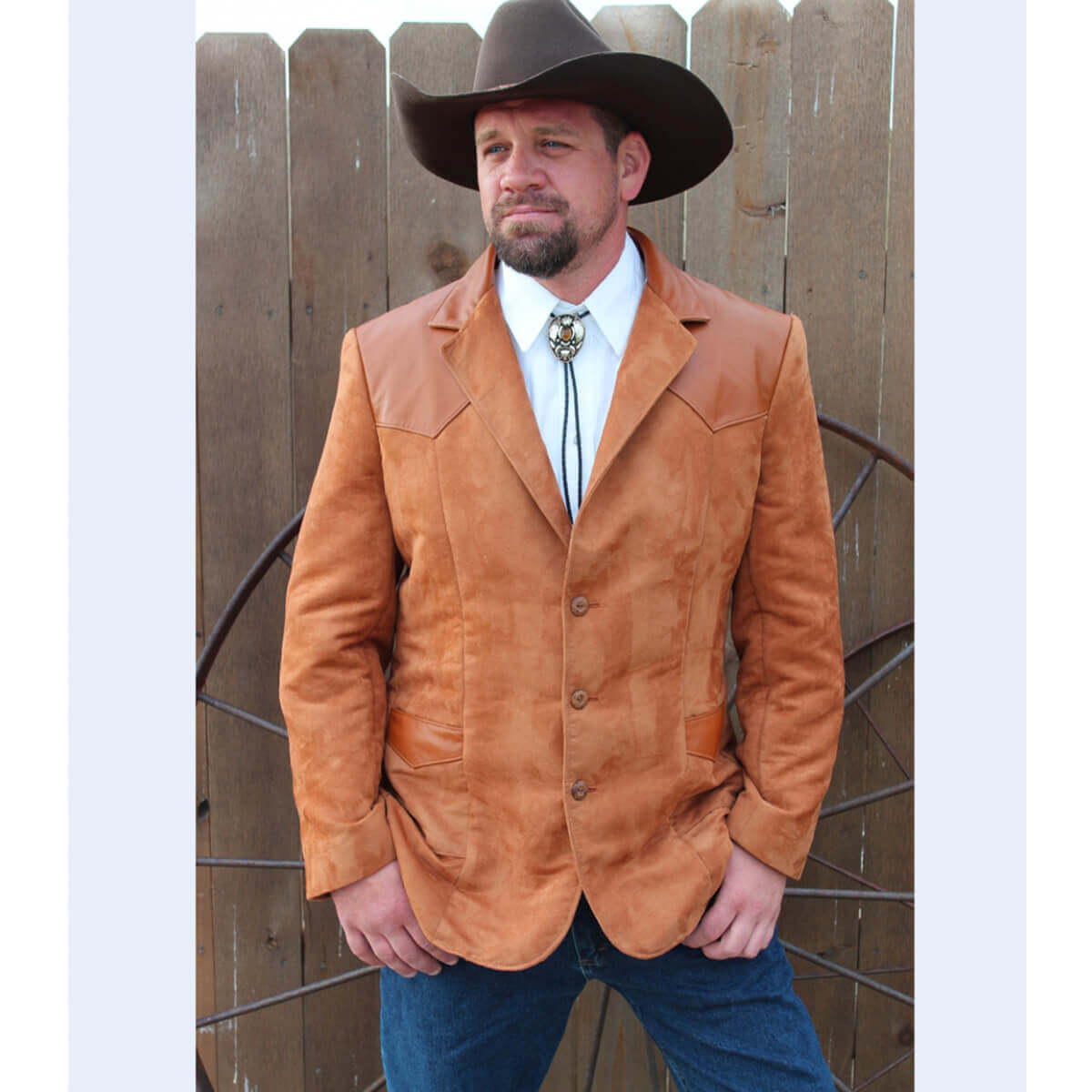 A man wearing a brown western blazer with contrasting black leather accents on the yokes, paired with a white shirt and blue jeans, standing in front of a wooden fence.
