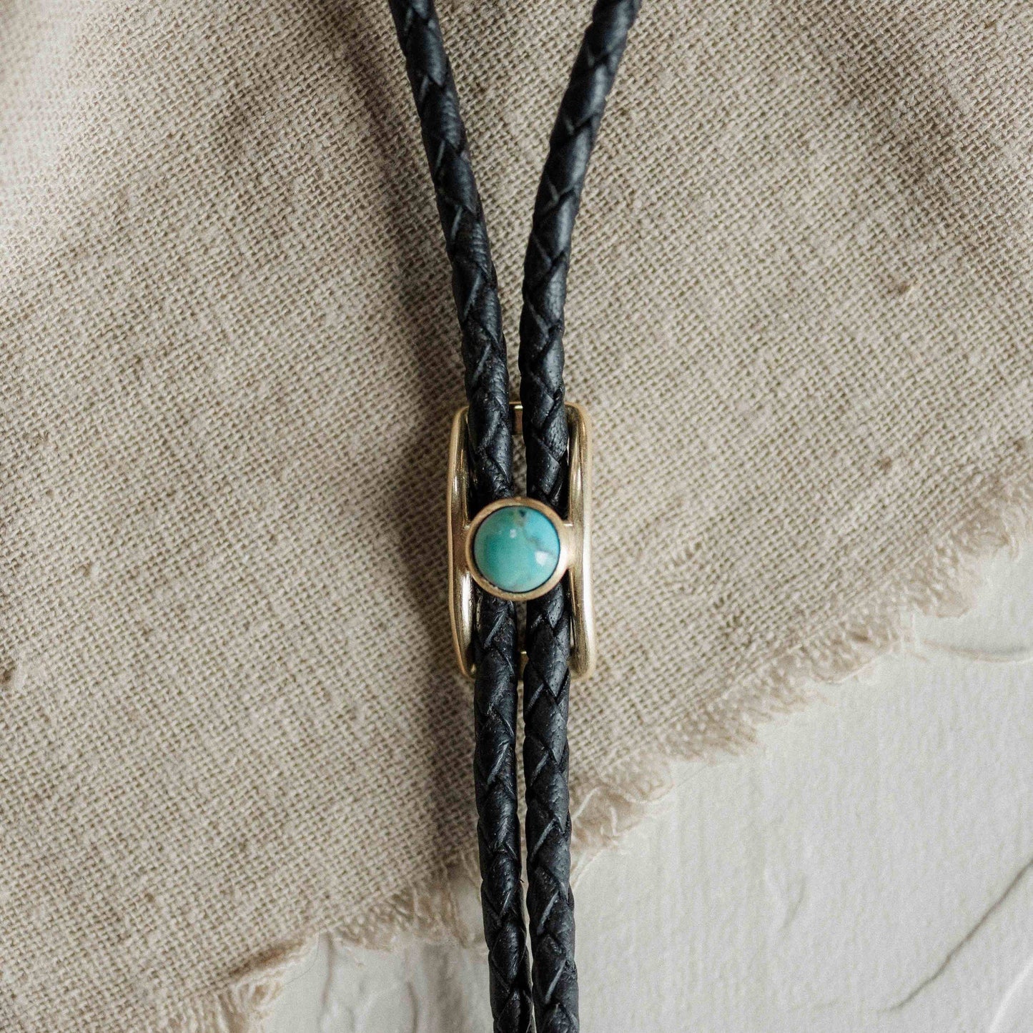 Black braided leather bolo tie with a turquoise stone on a textured beige background