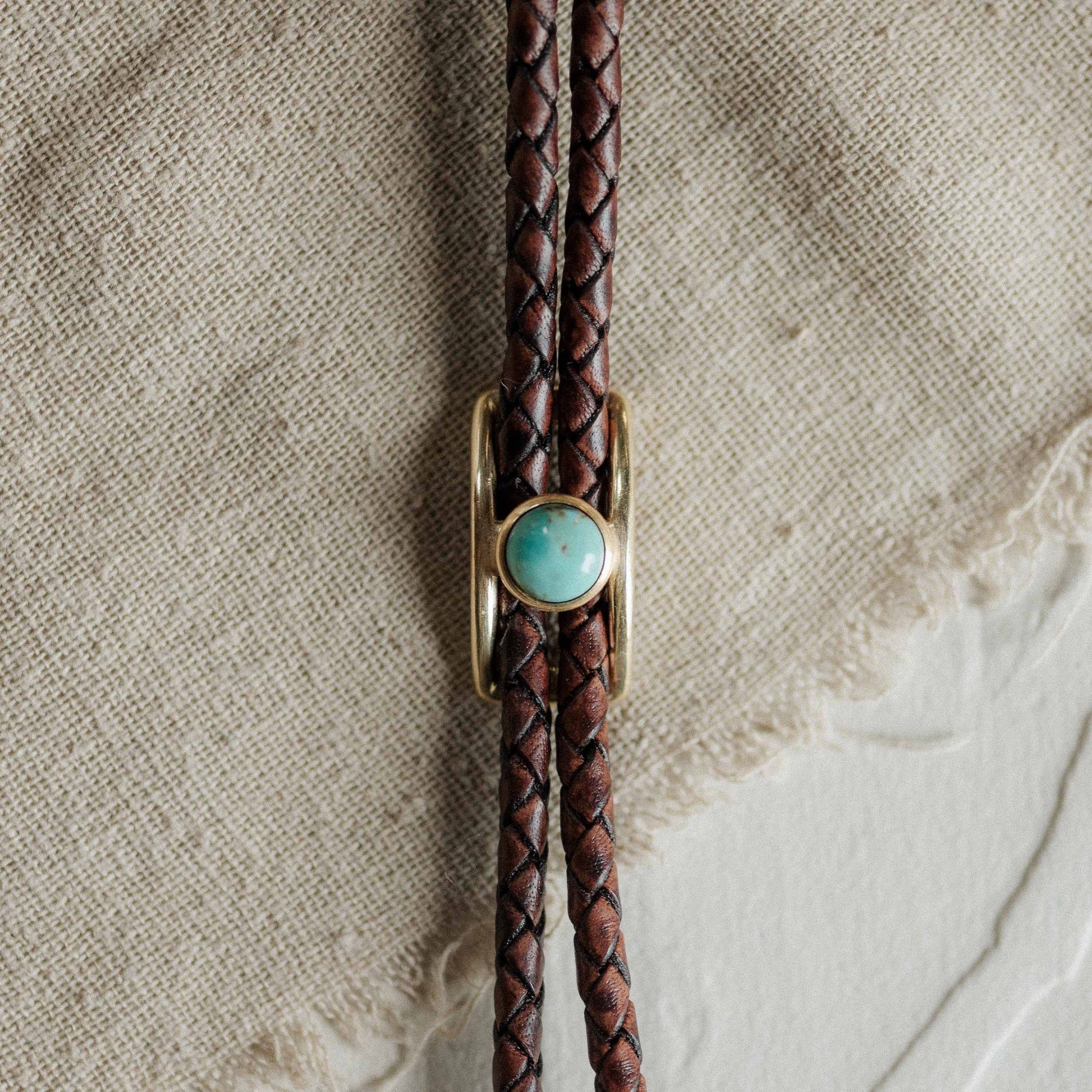 Brown braided leather bolo tie with a turquoise stone and gold clasp on a textured beige background.