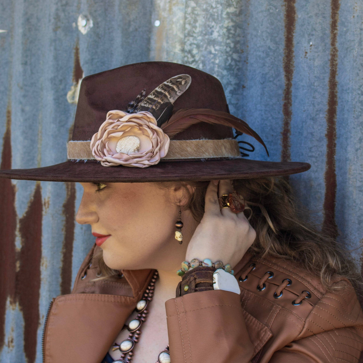 Mannequin wearing a brown hat with floral and feather details against a corrugated metal background.