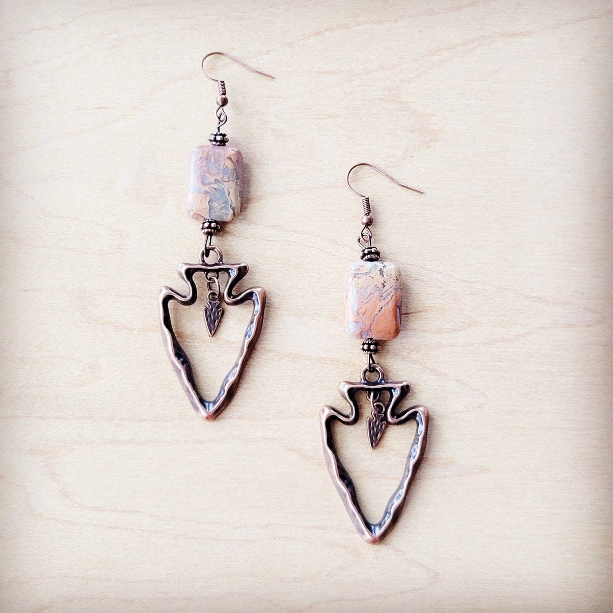 A pair of dangling earrings with textured bronze arrowhead charms, small marbled beads, and copper-toned hooks, displayed on a light wood background.