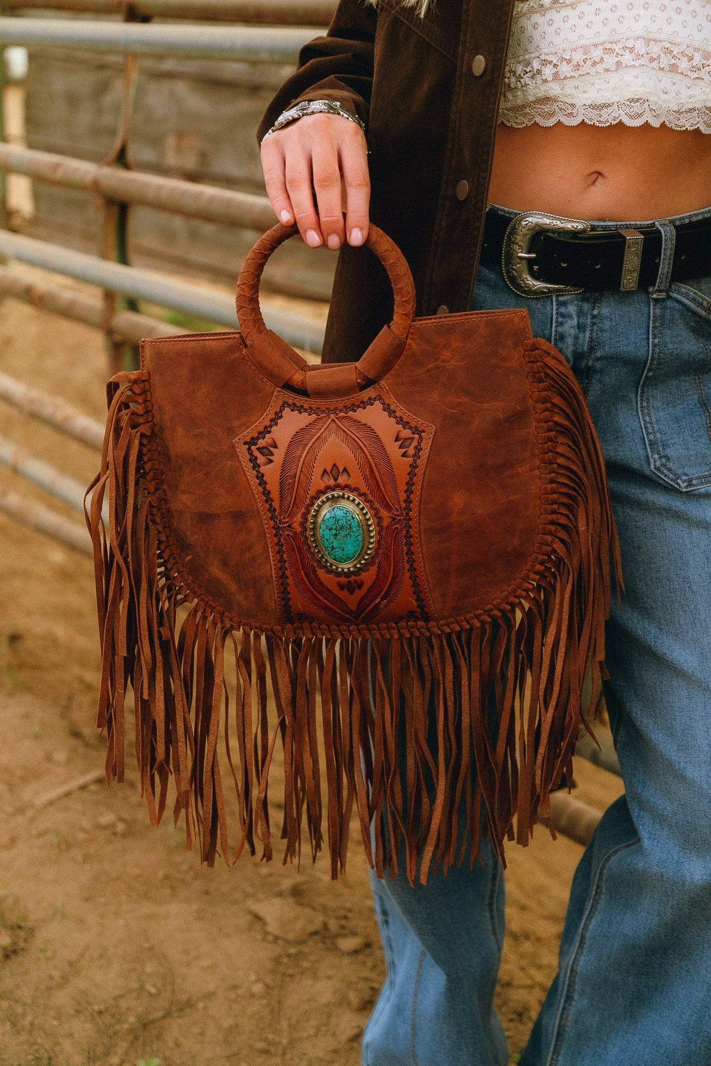Person holding a brown leather handbag with fringe and a turquoise stone, wearing jeans and a jacket.
