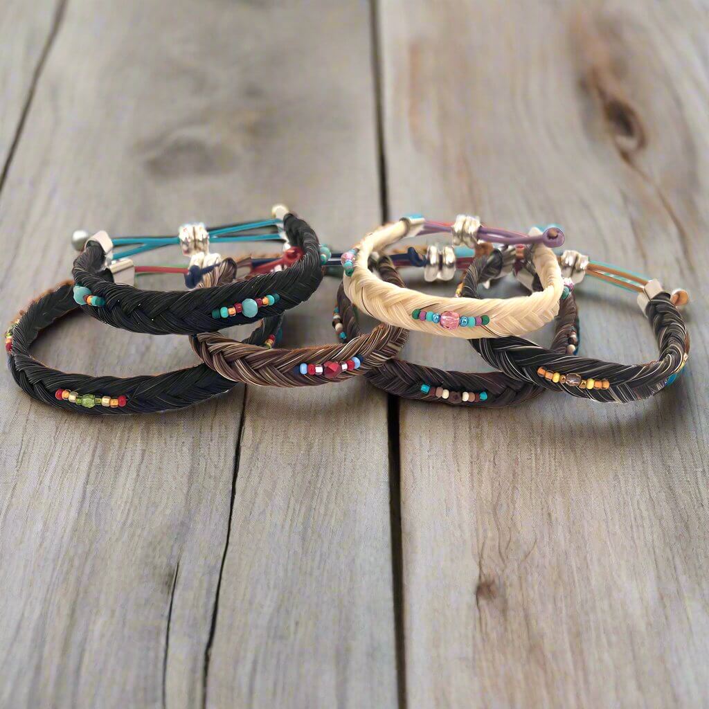 A set of five braided horsehair bracelets in black, brown, and beige, decorated with colorful seed beads and silver-toned metal spacers, arranged on a rustic wooden surface.