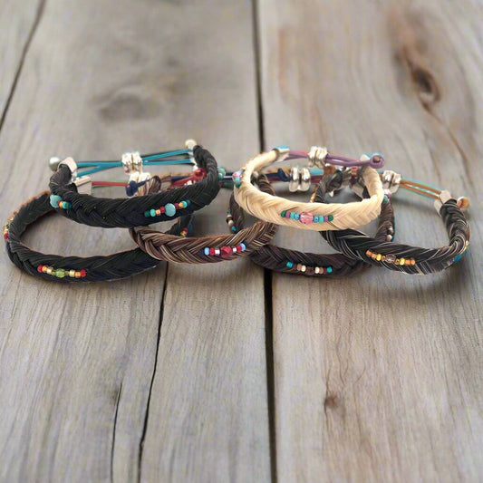 A set of five braided horsehair bracelets in black, brown, and beige, decorated with colorful seed beads and silver-toned metal spacers, arranged on a rustic wooden surface.