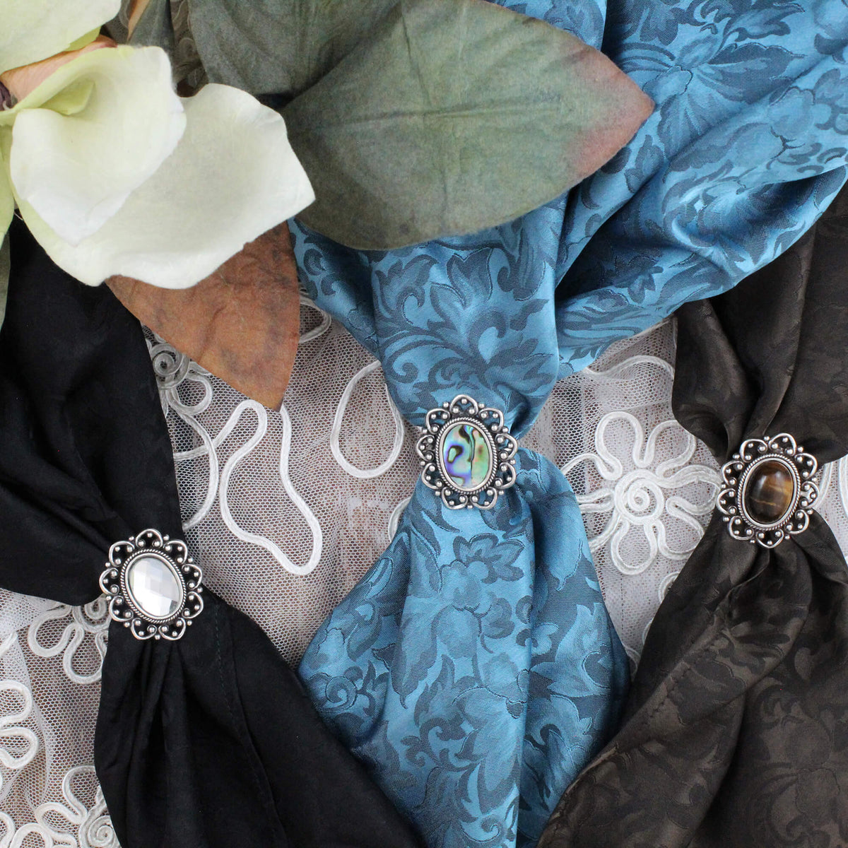 Three silk scarves in black, blue, and dark brown, each tied with a decorative silver-toned ring featuring an oval stone, arranged with green leaves on a white patterned fabric background.