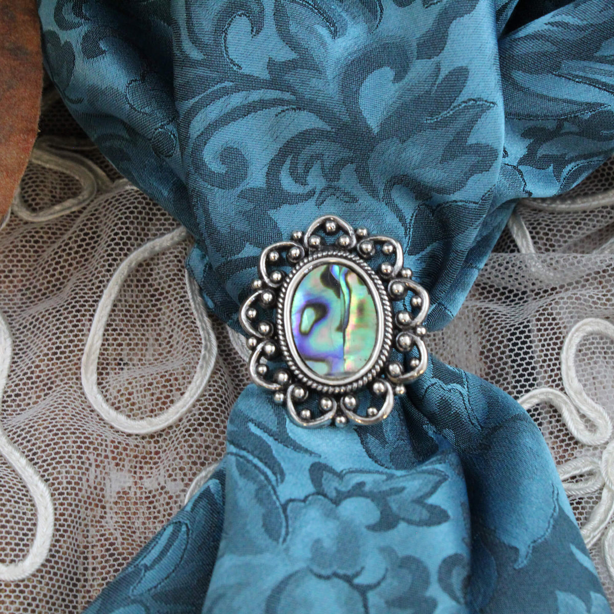 A close-up of a silver-toned ornate scarf slide with an iridescent abalone shell center, knotted through a blue silk scarf.