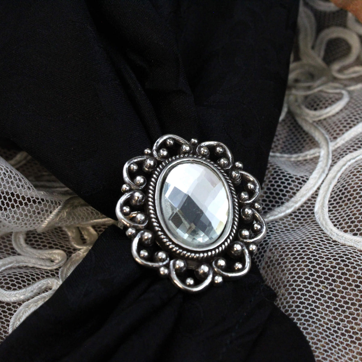 An ornate silver-toned scarf slide with a large oval clear faceted gemstone, displayed on a black scarf.