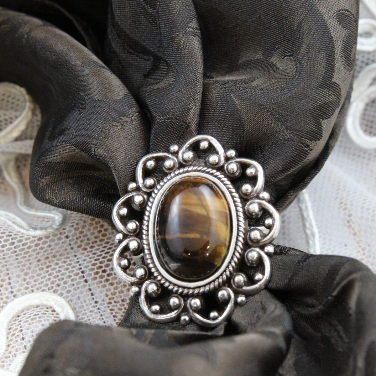A close-up of a silver-toned ornate scarf slide with an oval brown tiger's eye stone, holding a brown silk scarf.