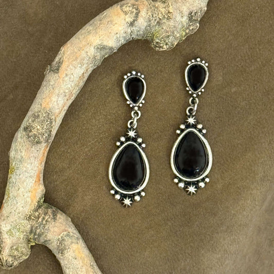 Black teardrop earrings with silver accents on a brown surface with a branch.