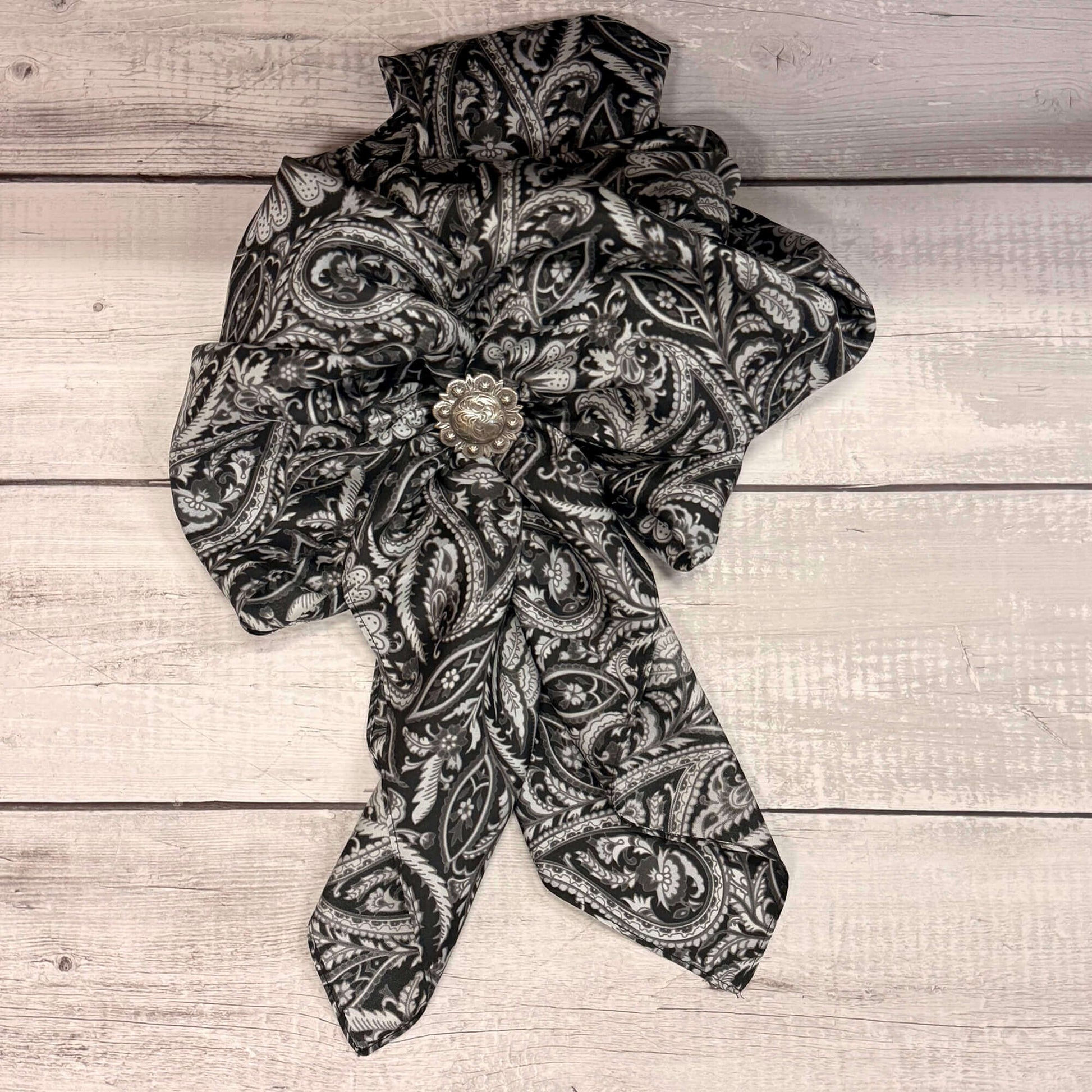 Black Paisley Silk Cowboy Western Wild Rag displayed on a wooden surface with a floral pin.