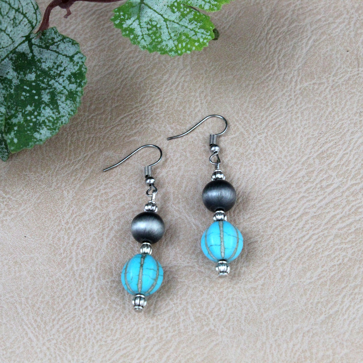 A pair of handmade dangle earrings featuring turquoise beads, dark gray round beads, and silver-toned metal spacers and hooks, displayed on a textured beige surface with green leaves in the corner.
