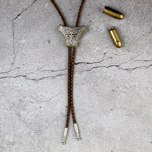 A brown braided rope bolo tie with a silver-toned metal shield-shaped concho featuring boot stitch design displayed on a textured gray concrete surface next to two brass bullets.