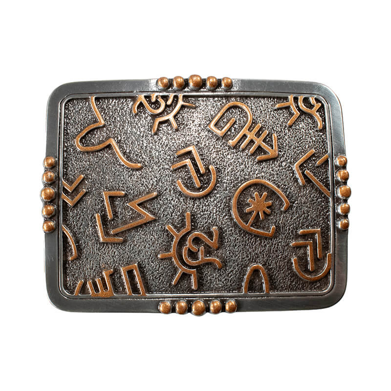 A rectangular metal belt buckle featuring a textured dark gray center with raised copper-colored indigenous-style symbols, surrounded by a dark metal border with small copper bead accents at the corners.