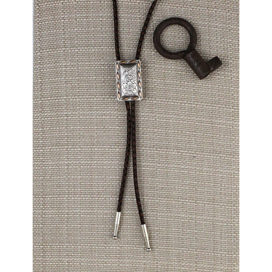 A western-style bolo tie with a silver-toned engraved rectangular center and dark brown braided cords, displayed on a textured beige fabric background next to a small dark metal key.